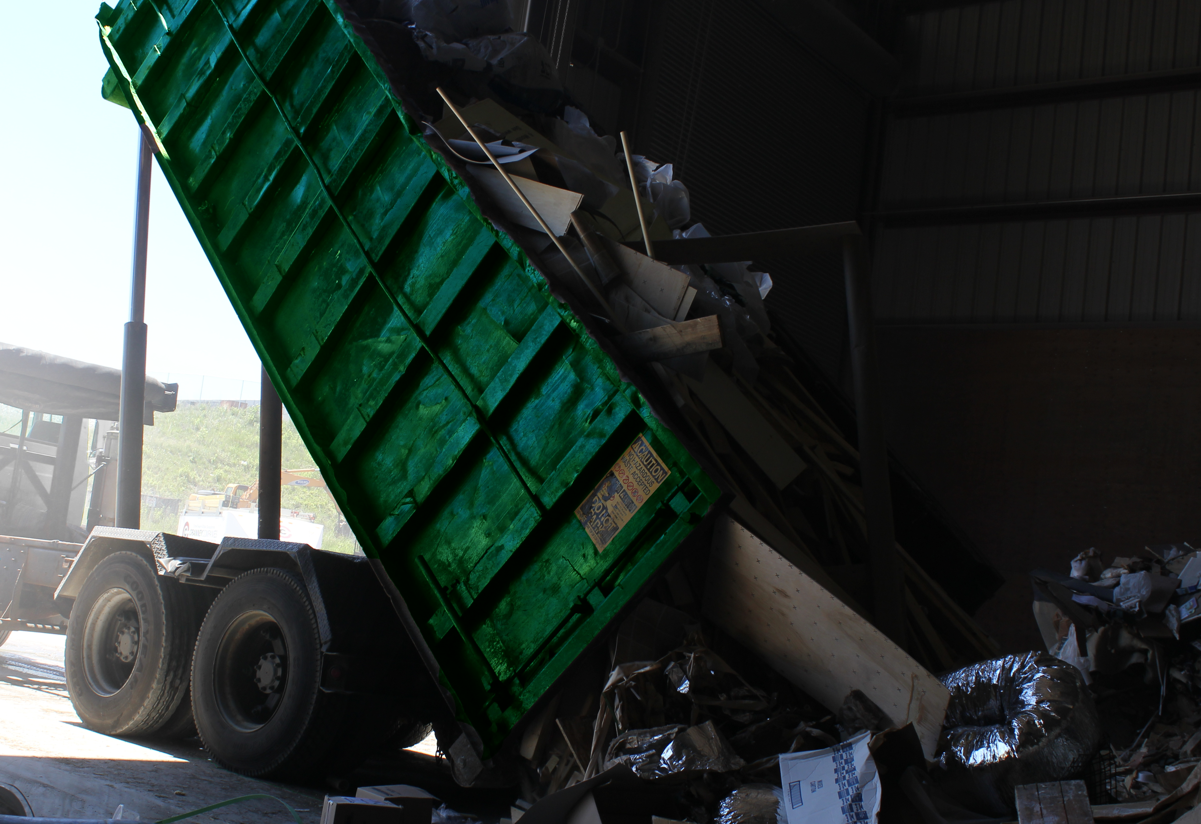 A green dumpster tilted upwards, unloading construction debris inside a dimly lit warehouse. Sunlight streams through, highlighting dust and debris.